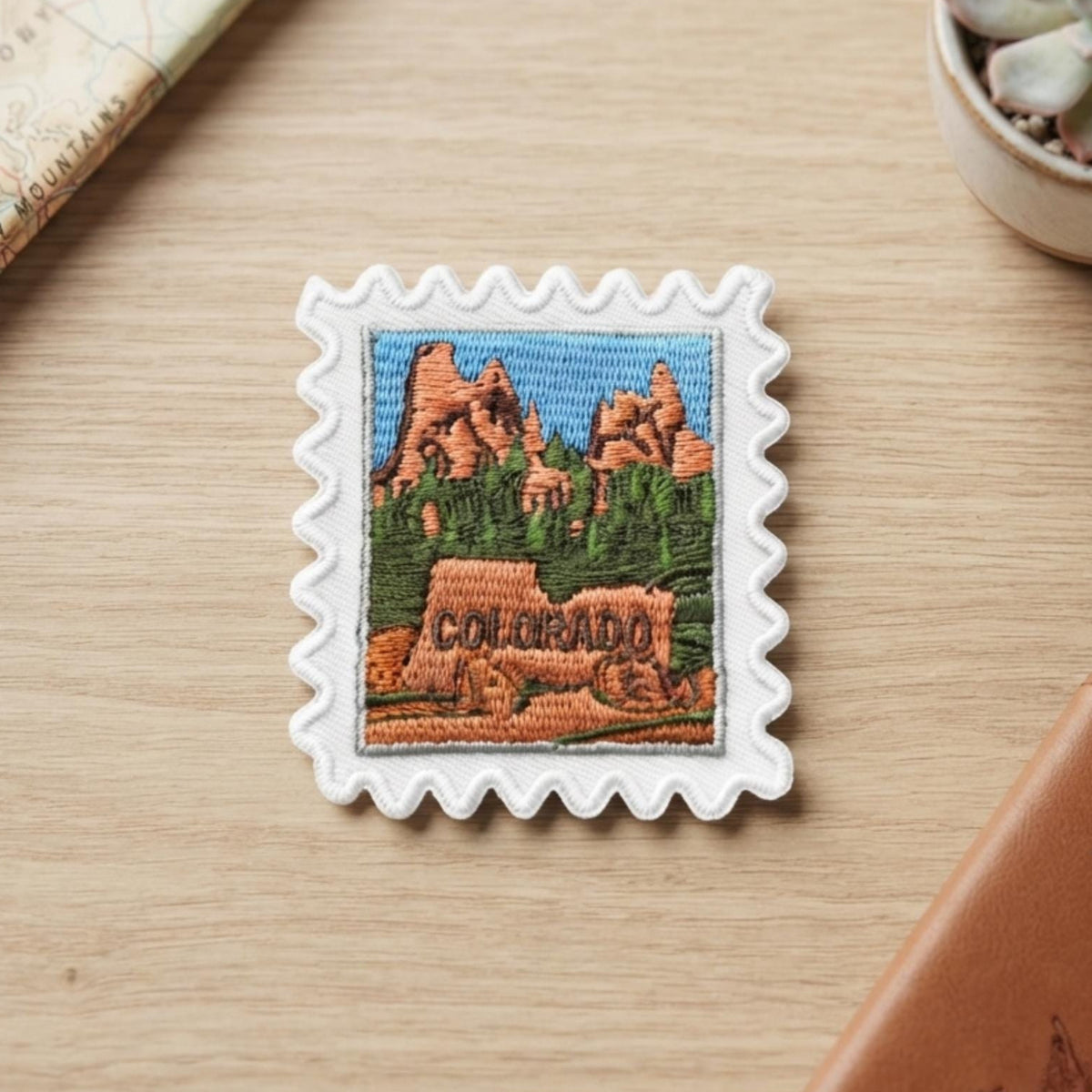 A square white embroidered patch with scalloped edges, featuring a colorful Colorado landscape and the word 'COLORADO' stitched in the foreground, displayed on a light wood tabletop with a passport, notebook, and succulent nearby.