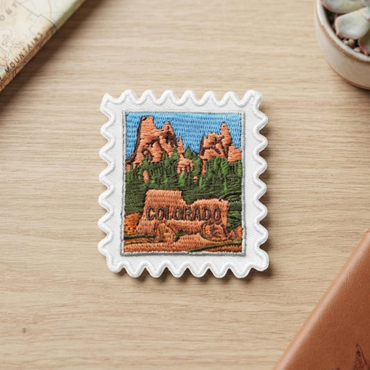 A square white embroidered patch with scalloped edges, featuring a colorful Colorado landscape and the word 'COLORADO' stitched in the foreground, displayed on a light wood tabletop with a passport, notebook, and succulent nearby.